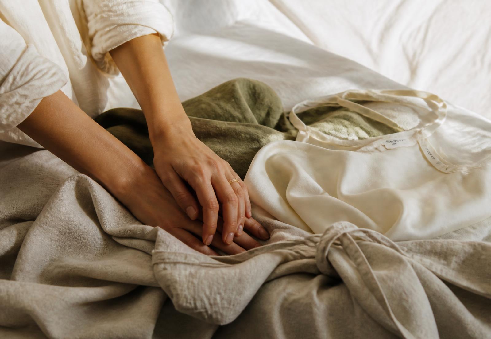 Hands resting on soft linen and silk garments laid across a bed